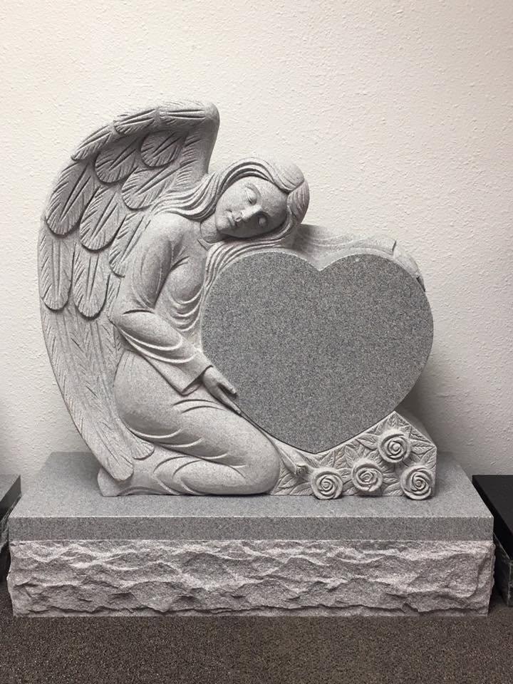 Gray granite sculpture monument featuring a kneeling angel with large detailed feathered wings resting her head tenderly on a large polished heart-shaped blank panel, with carved rose buds at the base, mounted on a rough rock-face base, displayed indoors in the Serenity Stone Co. showroom