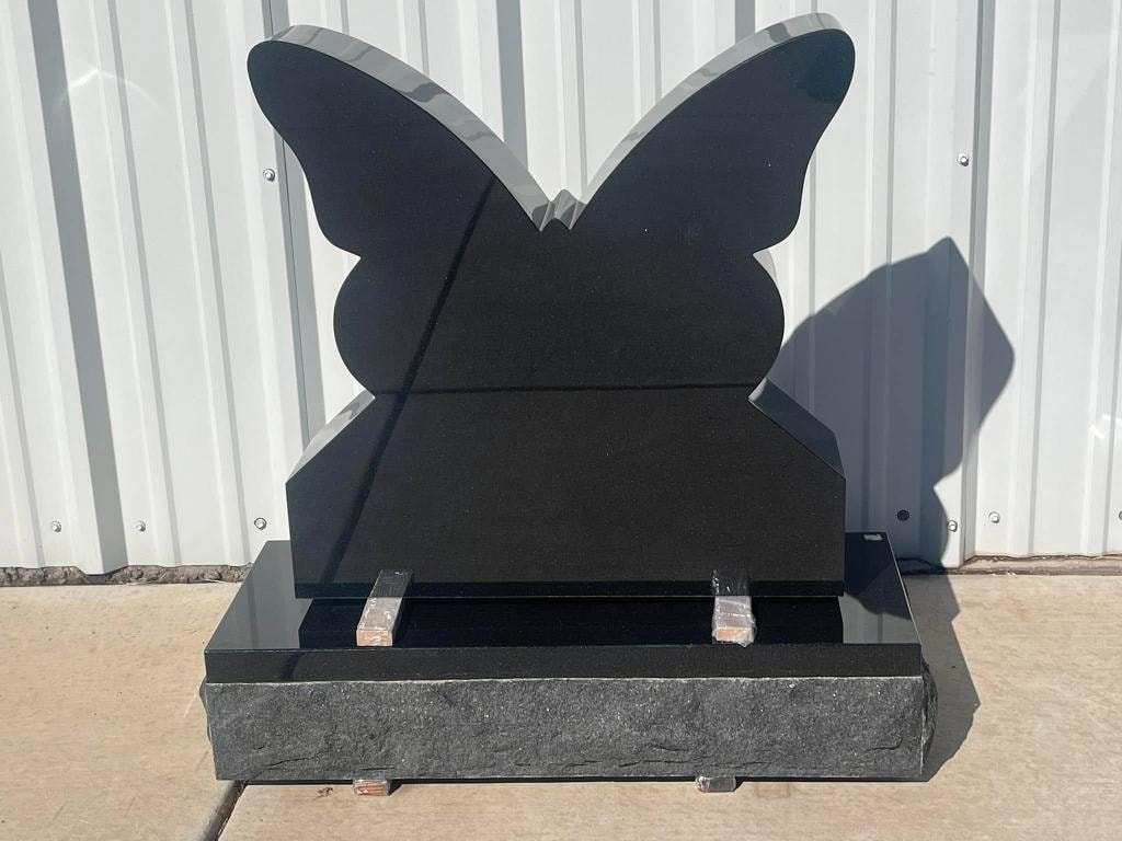 Polished black granite butterfly-shaped monument blank with beveled wing edges and rough-cut base, displayed outdoors against a metal building wall, ready for custom engraving