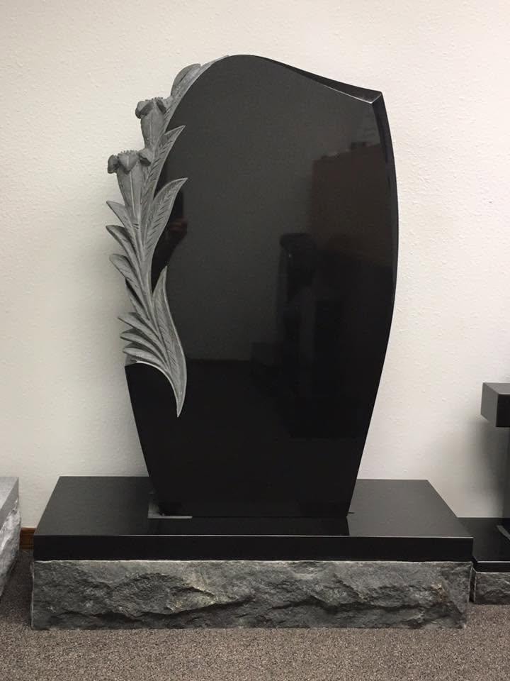 Polished black granite shield-shaped upright monument blank with a deeply carved three-dimensional carnation flower and long leafy stem running up the left side, mounted on a rough rock-face gray granite base, displayed indoors in the Serenity Stone Co. showroom, ready for custom engraving