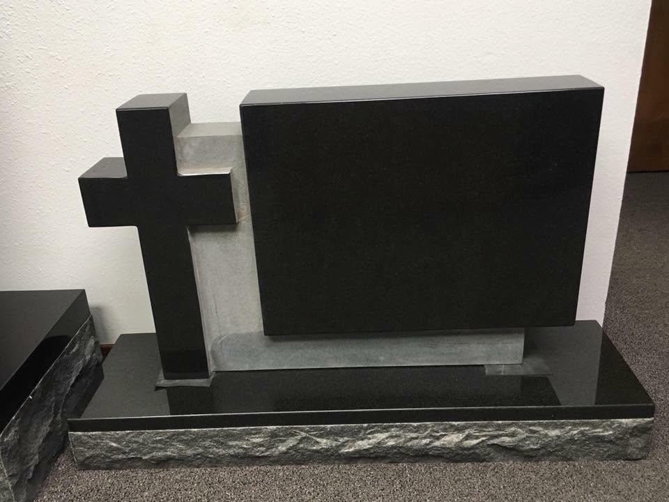 Polished black granite cross-and-tablet sculpture monument with a raised Latin cross on the left, large rectangular blank tablet panel on the right, and a rough rock-face base, displayed indoors in the Serenity Stone Co. showroom