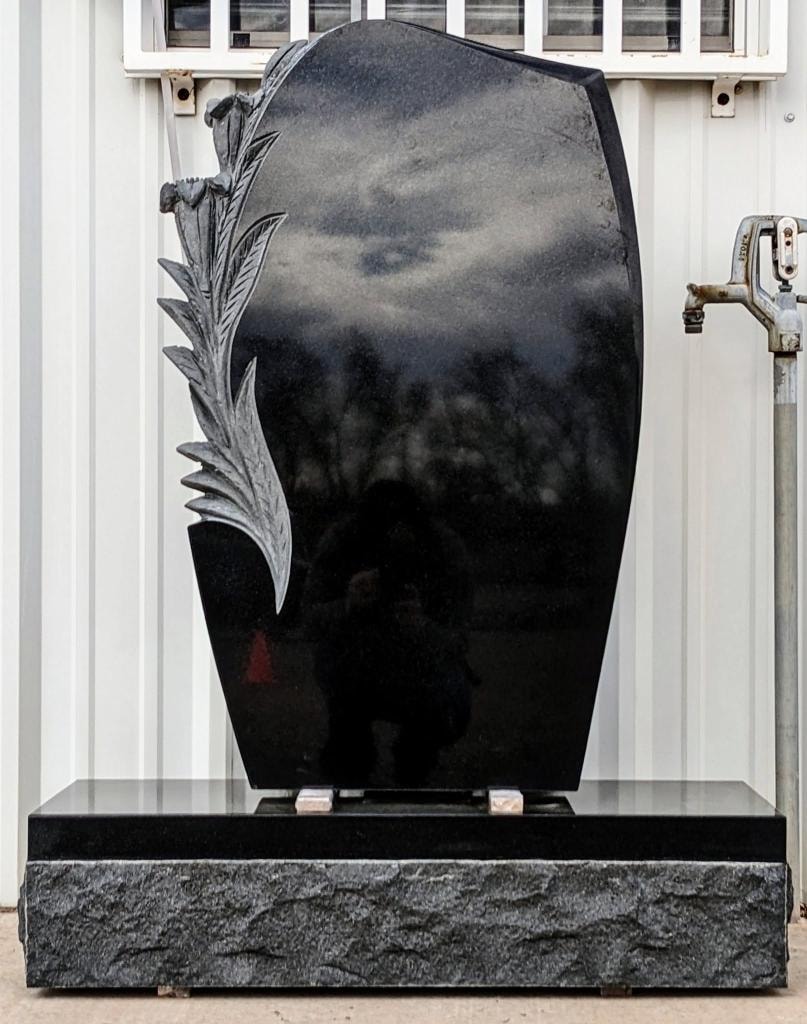 Polished black granite shield-shaped upright monument with a deeply carved three-dimensional feather and leaf spray sculpture on the left side, mounted on a rough-cut gray granite base, displayed outdoors against a white metal building