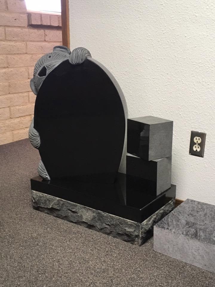 Polished black granite oval-shaped upright monument blank with deeply carved three-dimensional dolphins arching over the top, flanked by stacked polished black granite companion blocks on the right, mounted on a rough rock-face base, displayed indoors in the Serenity Stone Co. showroom