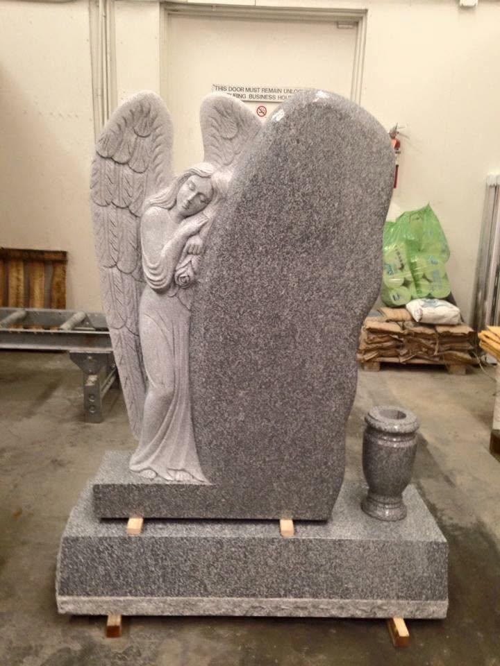 Gray granite upright monument blank with a deeply carved full-relief standing angel holding a rose, large outstretched wings framing the polished tablet panel, companion granite vase on the right, displayed on a two-tiered base indoors in the Serenity Stone Co. workshop
