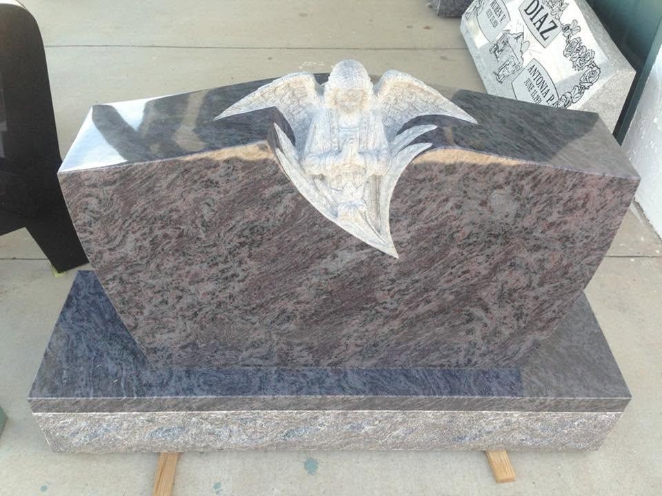Multicolor red and blue granite slant-style monument with a deeply carved three-dimensional praying angel emerging from a sculpted open book or wing shape at the center, mounted on a matching two-tiered granite base, displayed outdoors at the Serenity Stone Co. showroom