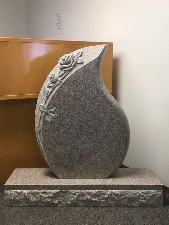 Gray granite teardrop-shaped monument with carved rose climbing the left edge, rough-cut base
