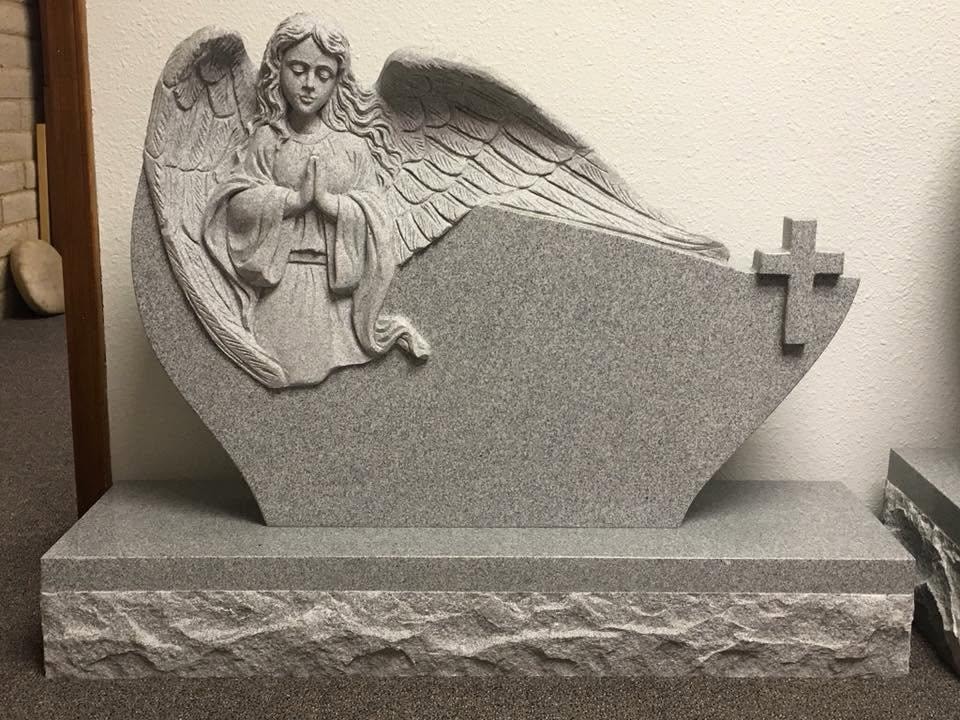 Gray granite monument blank with a deeply carved three-dimensional kneeling praying angel with outstretched wings on the left side and a raised cross on the right, polished center panel ready for custom engraving, mounted on a rough-cut rock-face base, displayed indoors in the Serenity Stone Co. workshop
