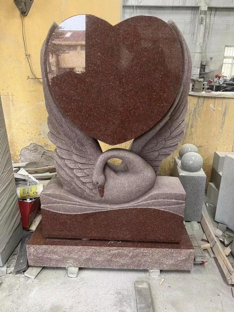 Red granite swan heart sculpture monument with a large polished heart top, carved swan with outstretched wings cradling the base, and a stepped rectangular base — displayed in a stone workshop