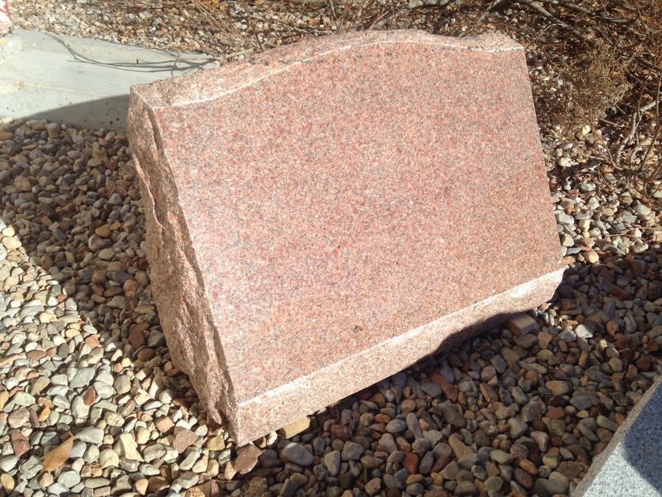 Polished red granite slant monument blank with rough-cut natural sides and base, displayed outdoors on gravel in the Serenity Stone Co. yard, ready for custom engraving