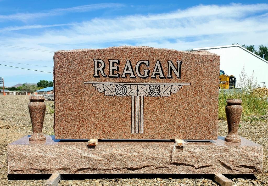 Reagan pink granite single upright monument with decorative column and floral engraving, companion vases, and rough-cut base in display yard