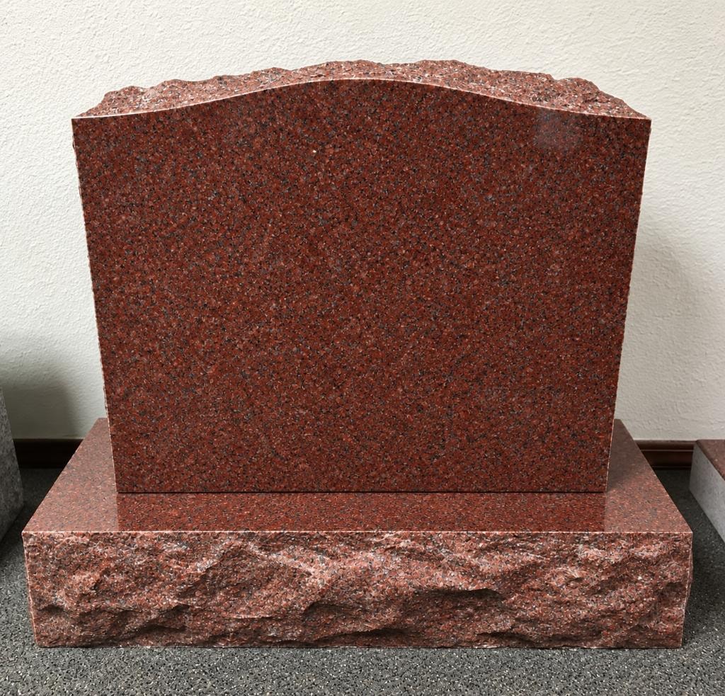 Polished red granite upright monument blank with rough-cut natural top edge and textured rock-face base, displayed indoors against a white wall, ready for custom engraving