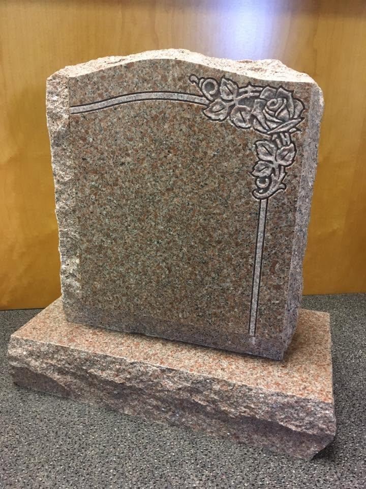 Pink granite upright monument blank with rough-cut sides and base, featuring an engraved long-stem rose with buds and leaves design on the upper right corner, displayed indoors